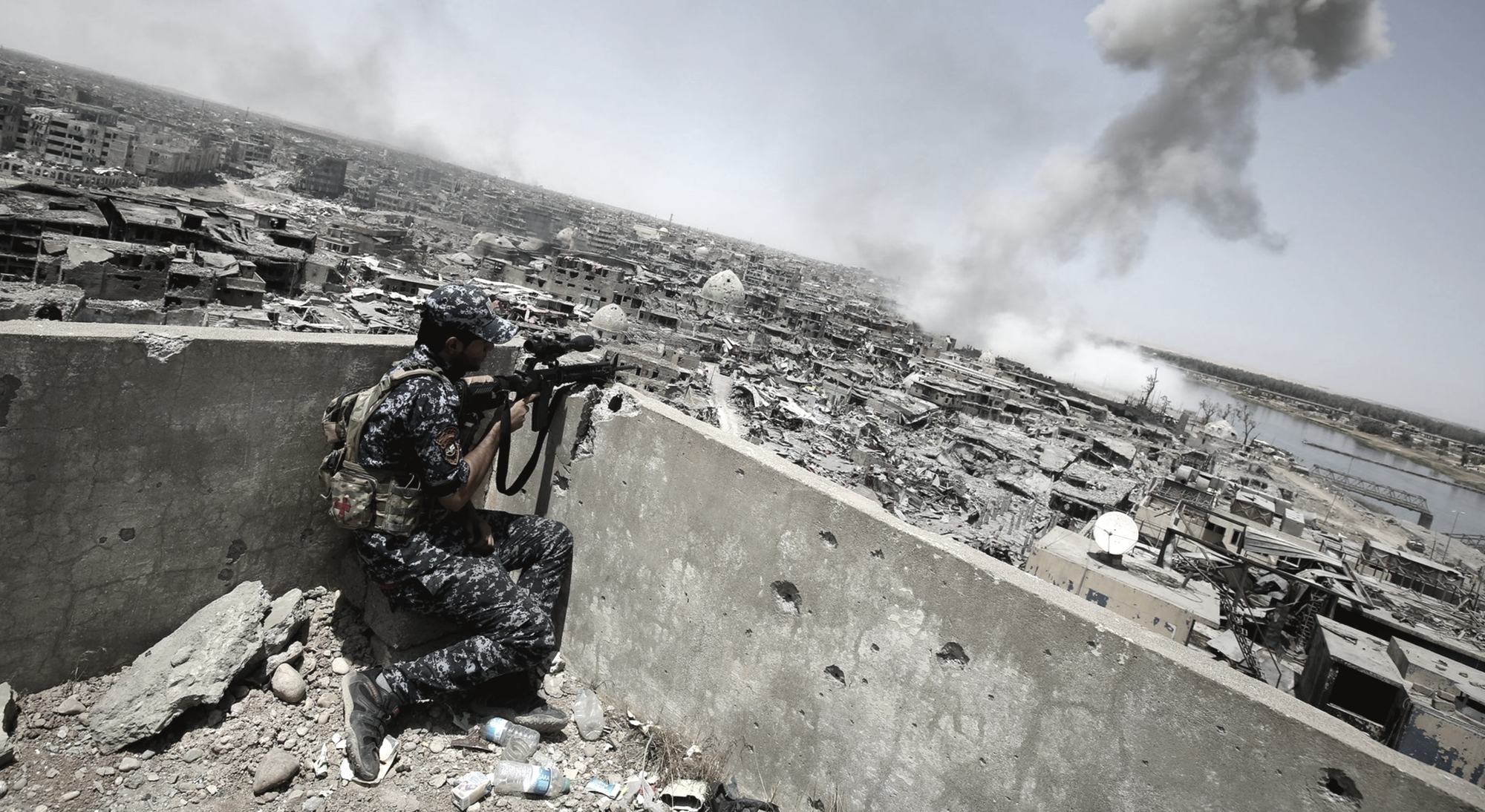 An Iraqi Federal Police sniper on a rooftop parapet above the ruined Old City of Mosul, the Tigris beyond and a smoke column rising