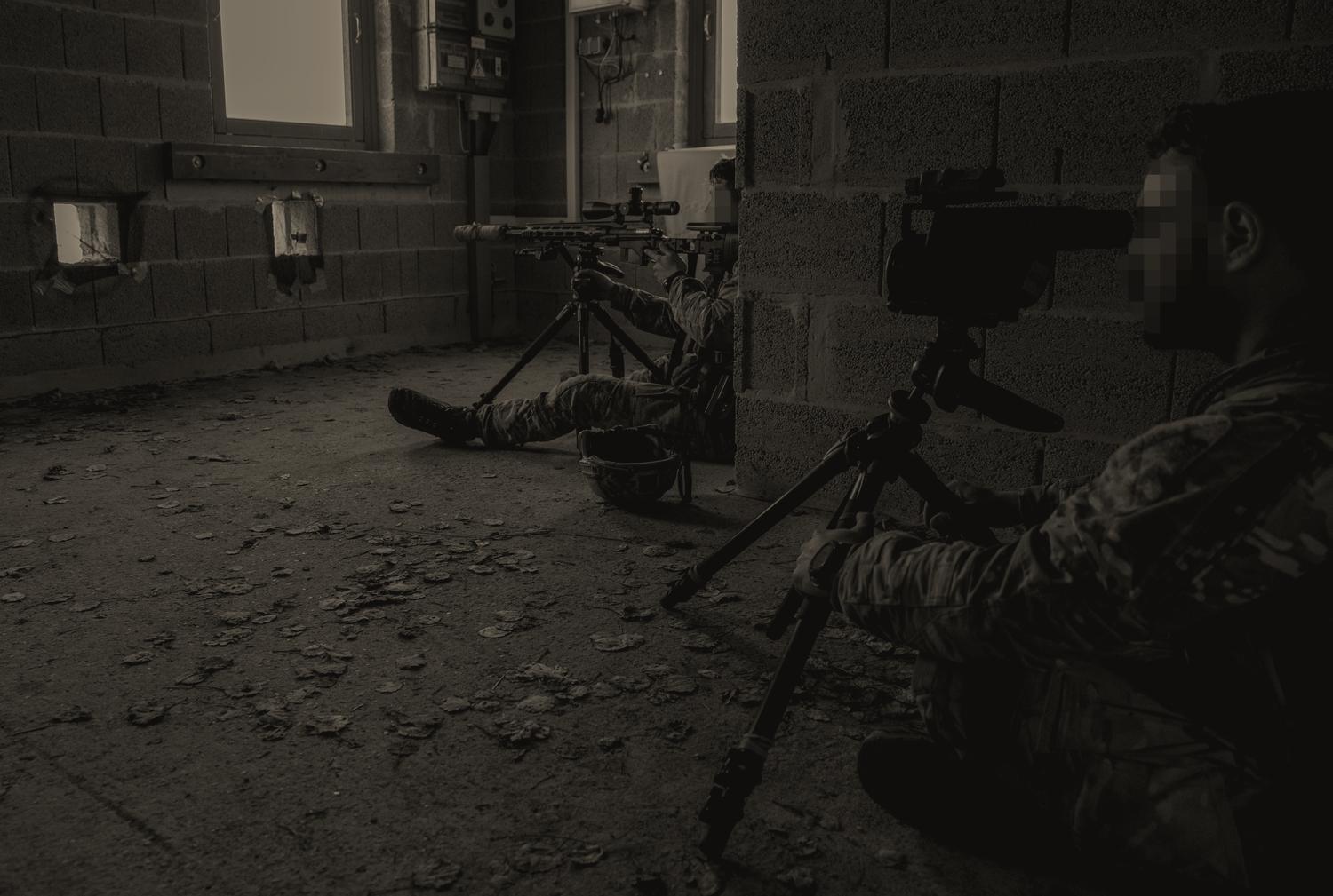 A sniper team in a breeze-block room on the International Special Training Center’s urban sniper course: one man behind a rifle on a tripod at a loophole cut in the wall, another behind a spotting scope