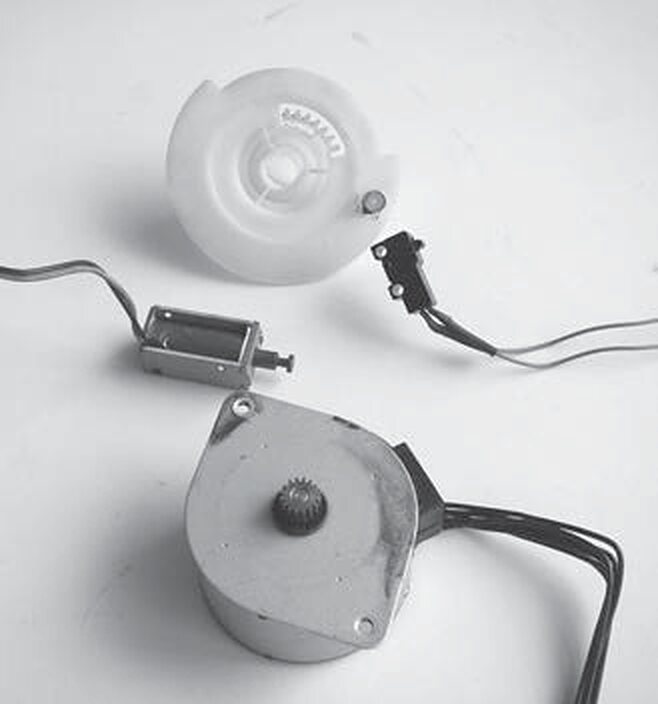 A stepper motor, the silver cylinder with its bundle of wires, beside a small linear actuator, a microswitch, and a white cam wheel